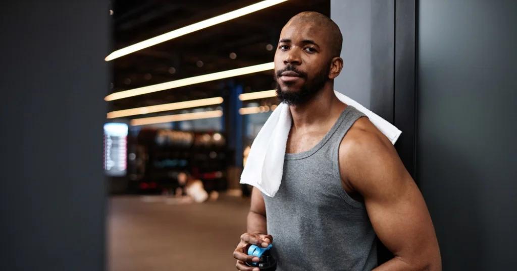 Fitness Coach in Birmingham relaxing post-workout with towel and drink, leaning against a wall in a modern gym setting.