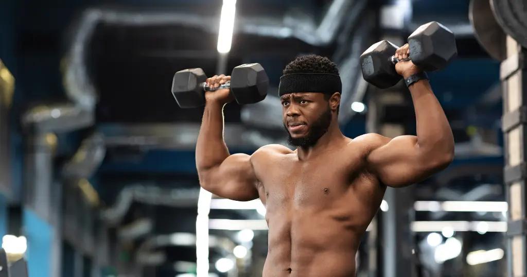 A gym trainer in Birmingham performing a shoulder press with dumbbells in a well-equipped gym.