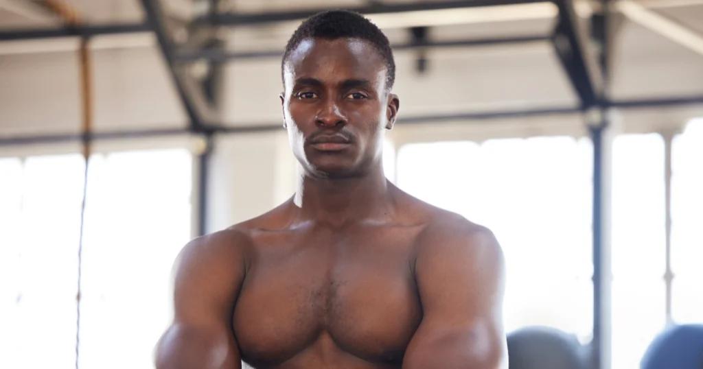 Confident Fitness Coach in Birmingham standing shirtless in a bright gym, showcasing a strong, muscular physique.